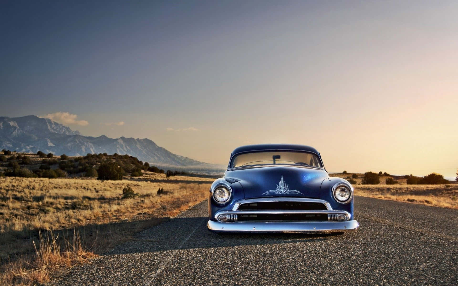 A Blue Classic Car Is Parked On A Road In The Mountains Background