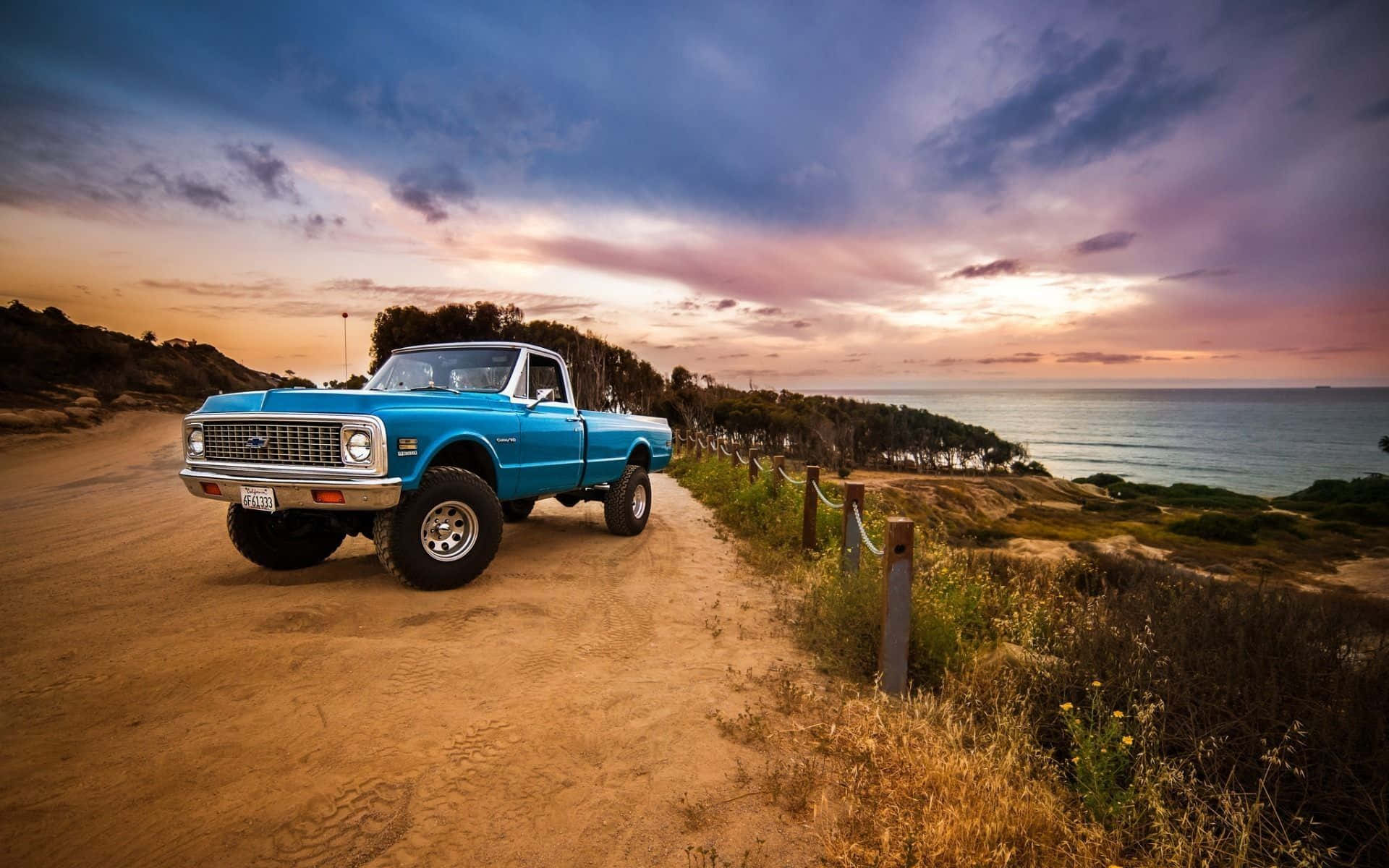 A Blue Truck Is Parked On A Dirt Road Near The Ocean Background