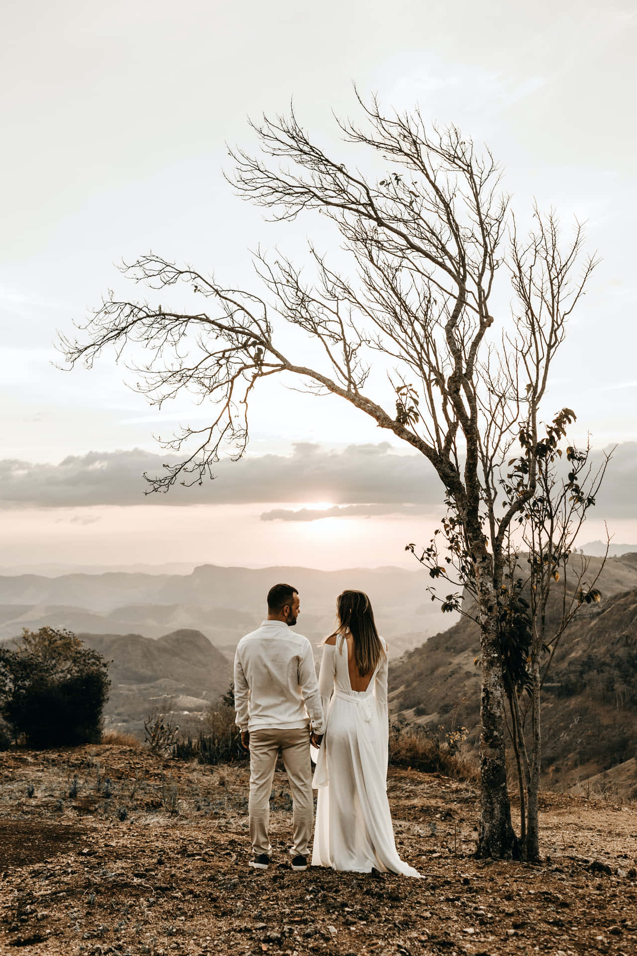 A Bride And Groom Standing On A Hill Overlooking The Mountains Background