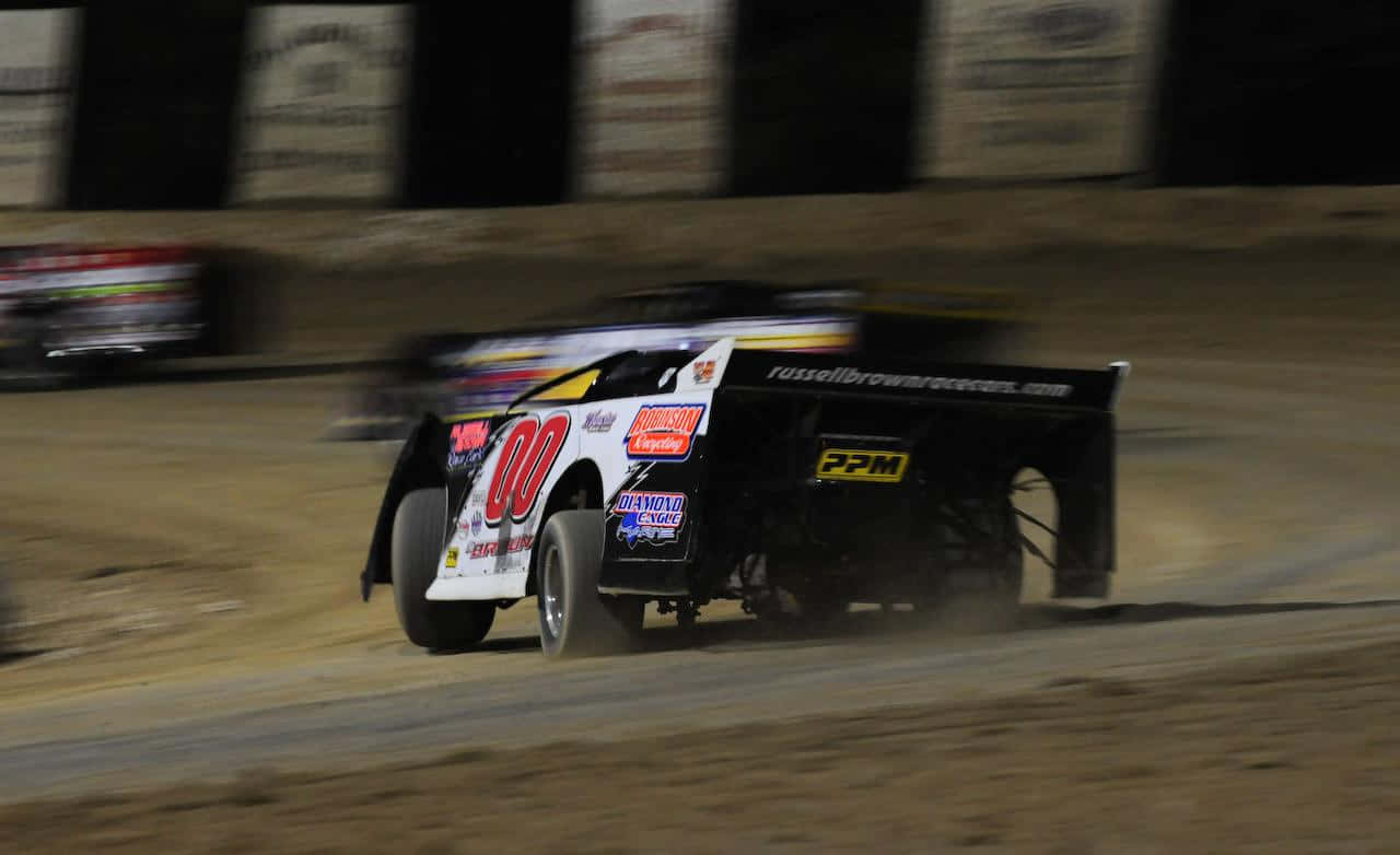 A Dirt Track With Several Cars Racing At Night Background