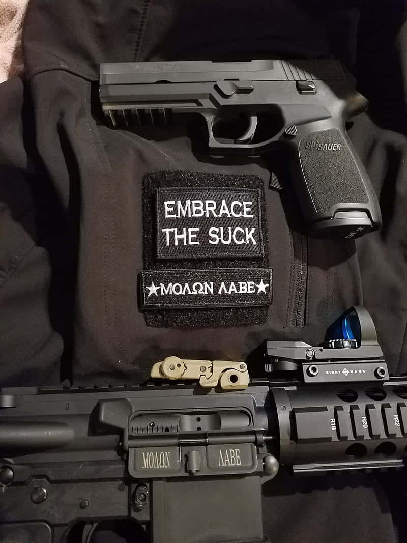 A Gun And A Patch With The Words Embrace The Suck Background