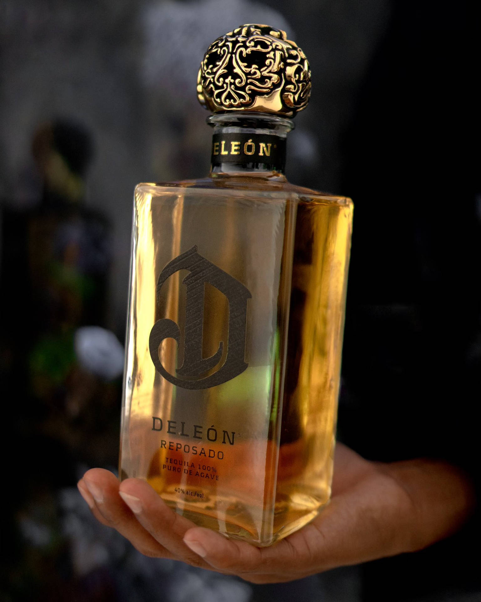 A Hand Holding A Bottle Of Deleon Tequila's Golden Reposado Background