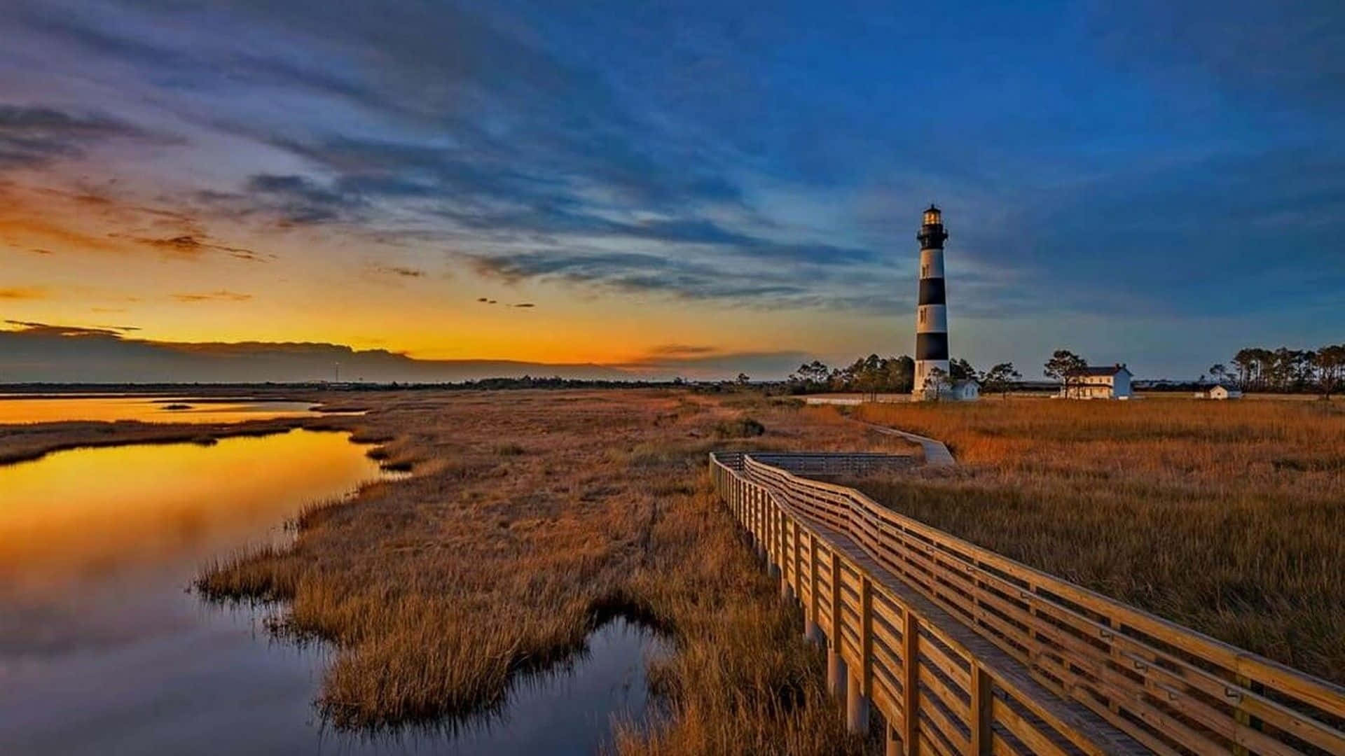 A Lighthouse Is Seen At Sunset Near A Marsh Background