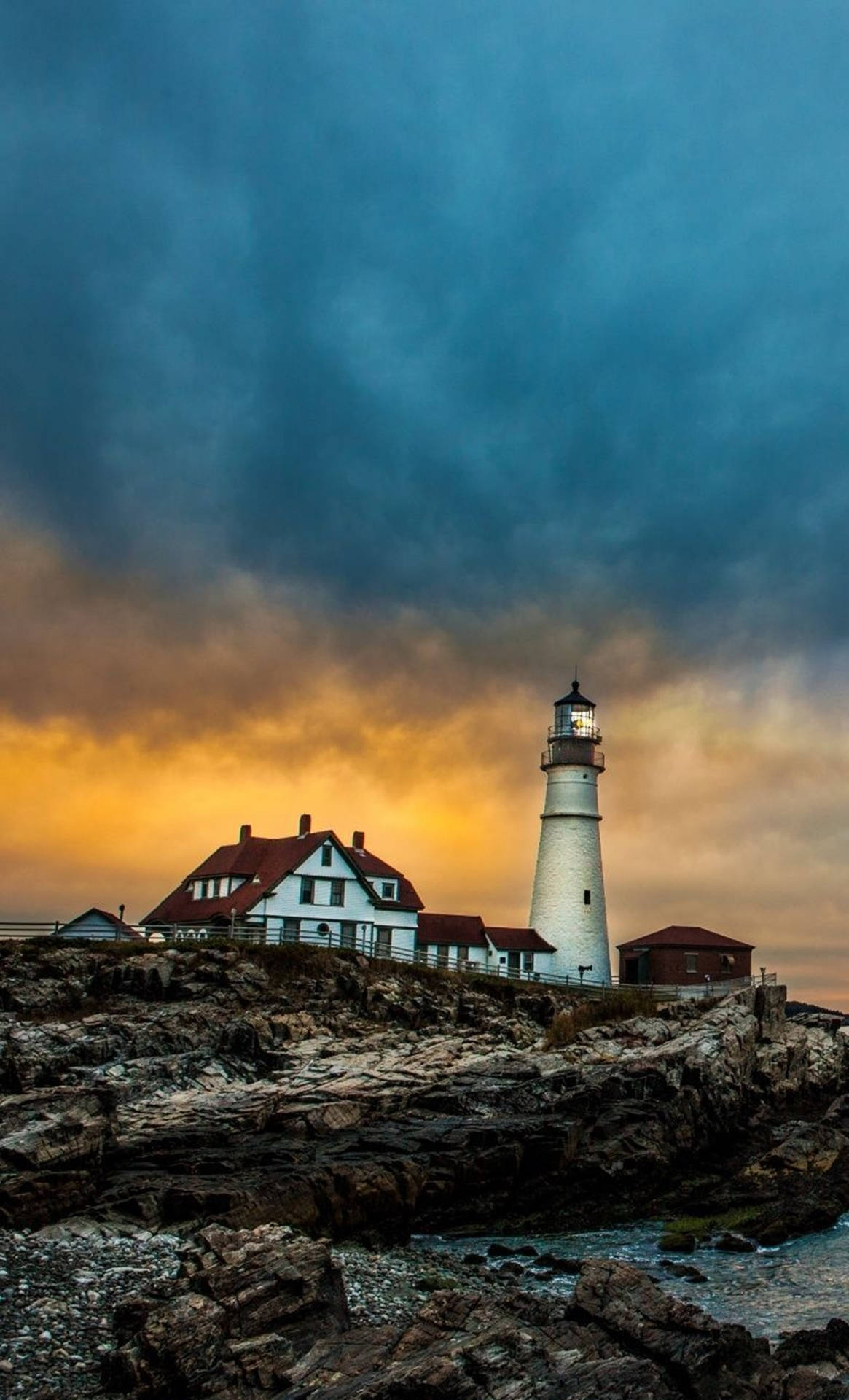 A Lighthouse Sits On A Rocky Shore With A Cloudy Sky Background