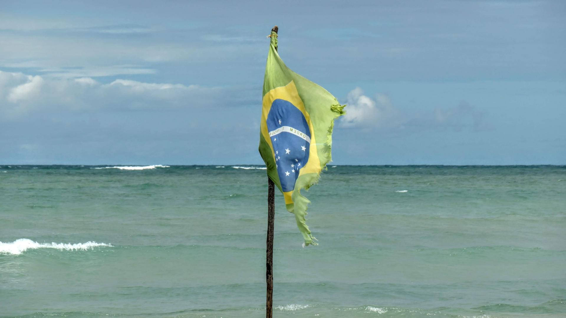 A Majestic Brazilian Flag Fluttering Against A Beautiful Beach Landscape Background