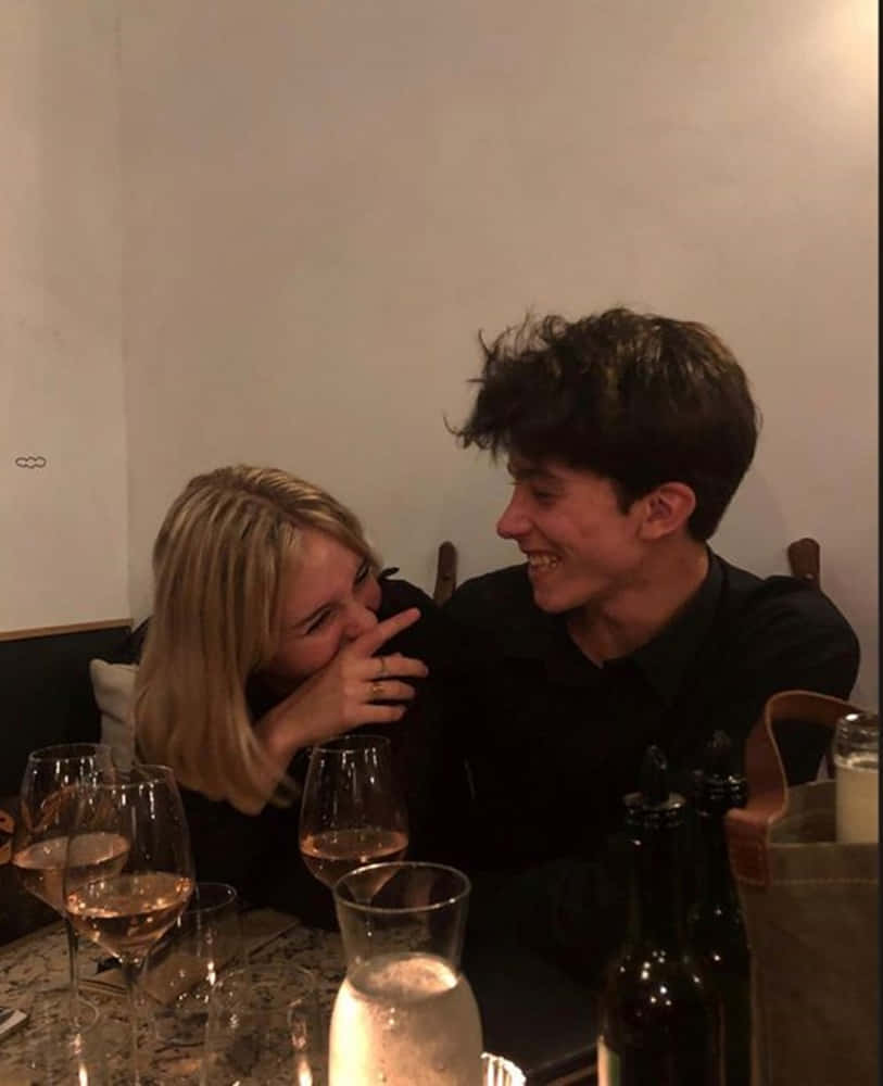 A Man And Woman Are Sitting At A Table With Wine Glasses Background