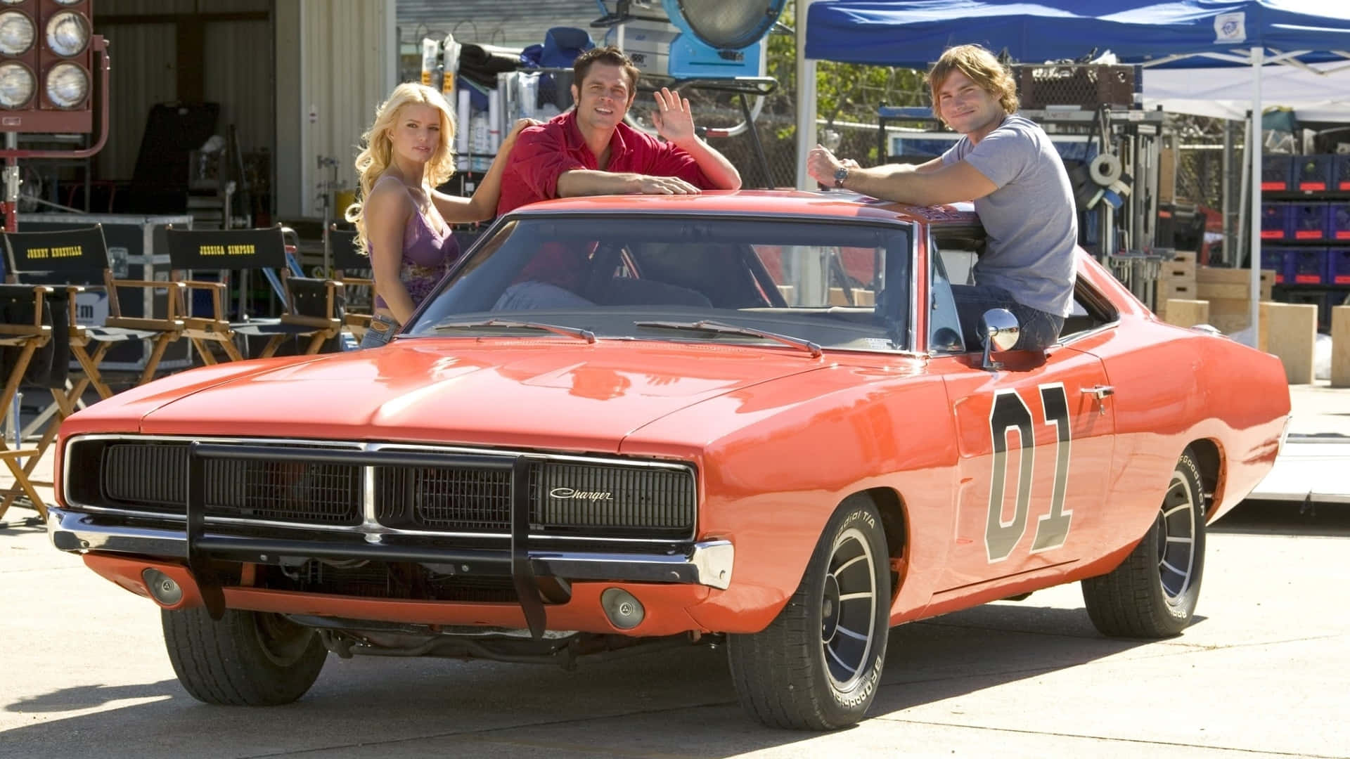 A Man And Woman Pose In Front Of An Orange Muscle Car Background