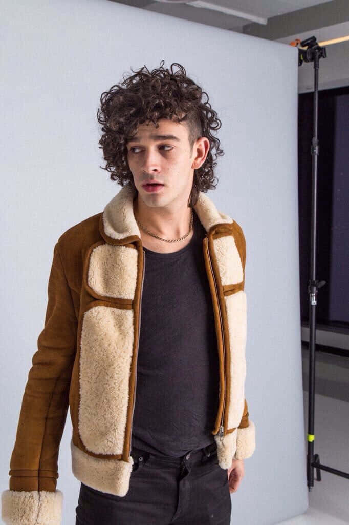 A Man In A Brown Shearling Jacket Standing In Front Of A Camera Background