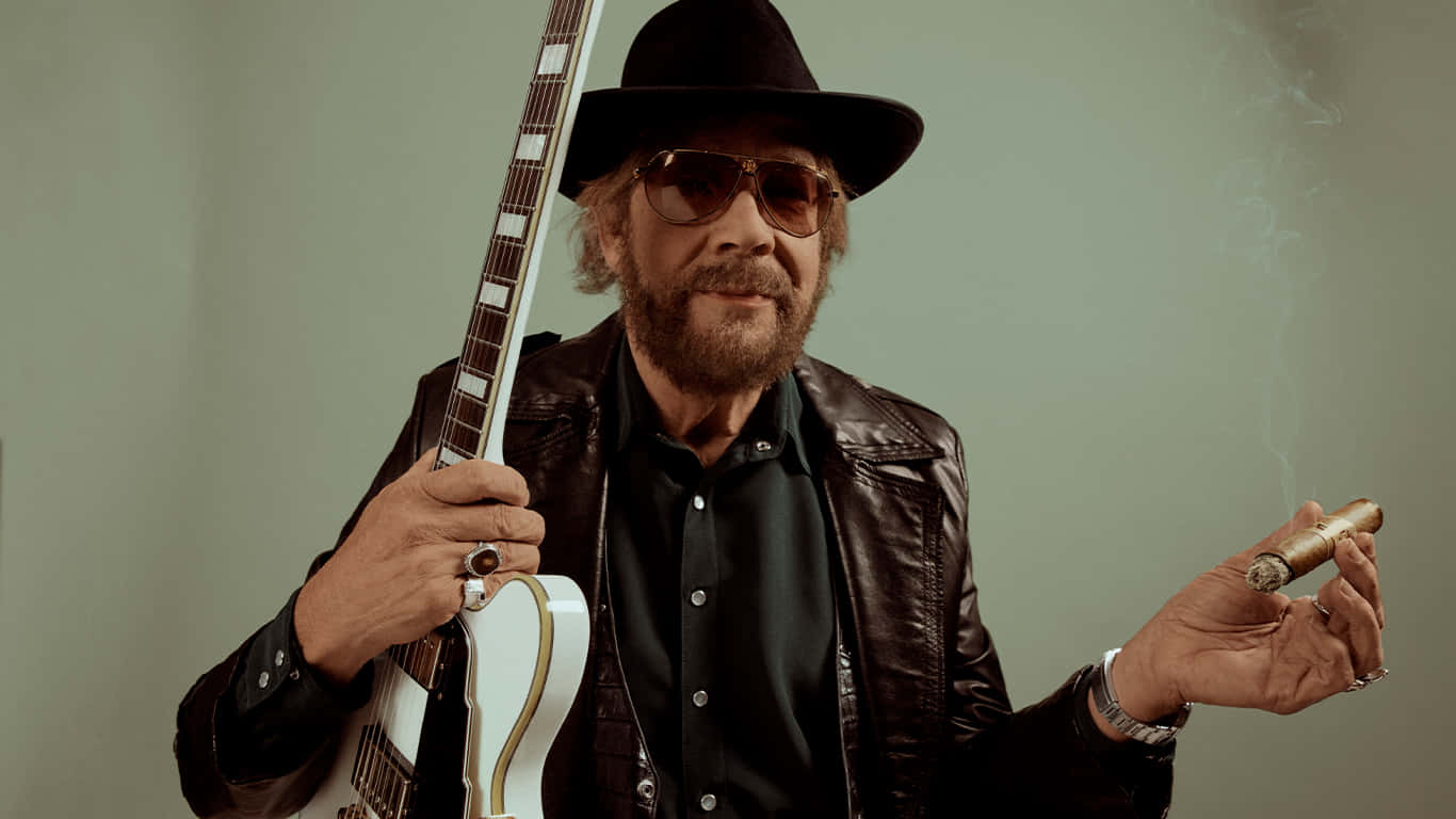 A Man In A Hat And A Jacket Holding A Guitar Background