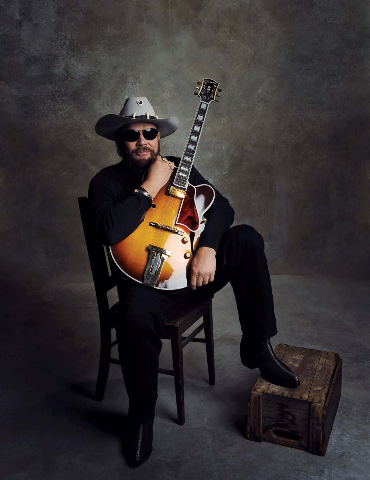 A Man In A Hat And Cowboy Hat Sitting On A Chair With A Guitar Background