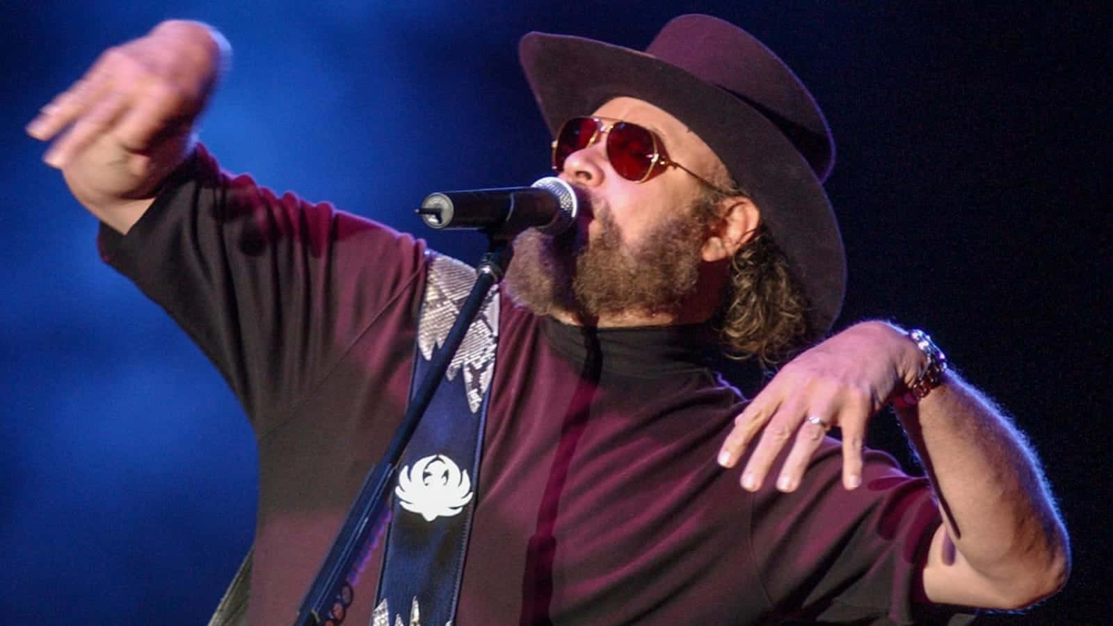 A Man In A Hat And Sunglasses Singing Into A Microphone