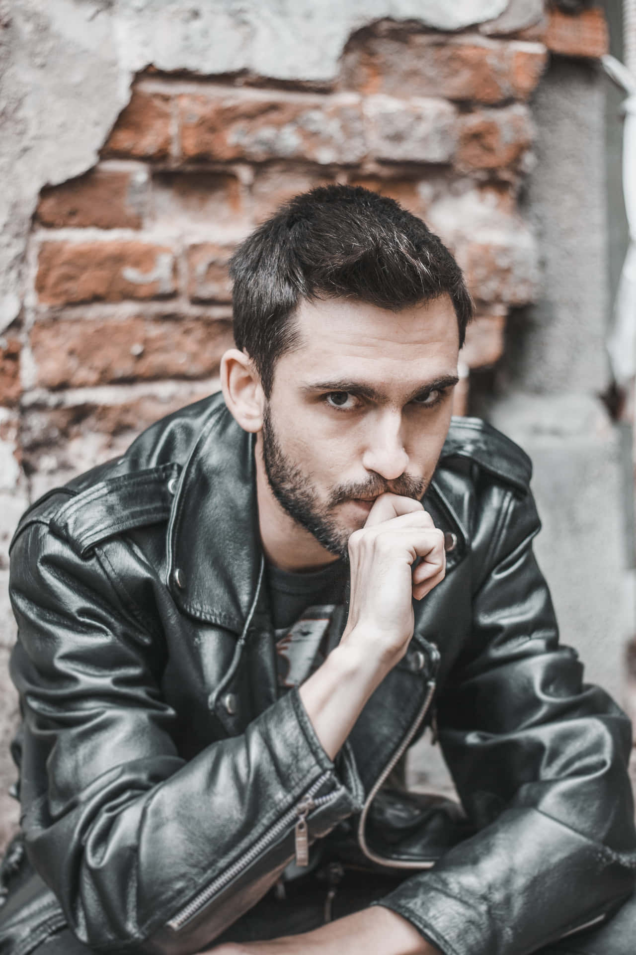 A Man In A Leather Jacket Is Sitting On A Brick Wall Background