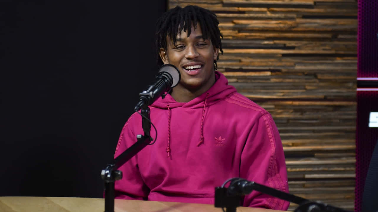 A Man In A Pink Hoodie Is Sitting In Front Of A Microphone Background