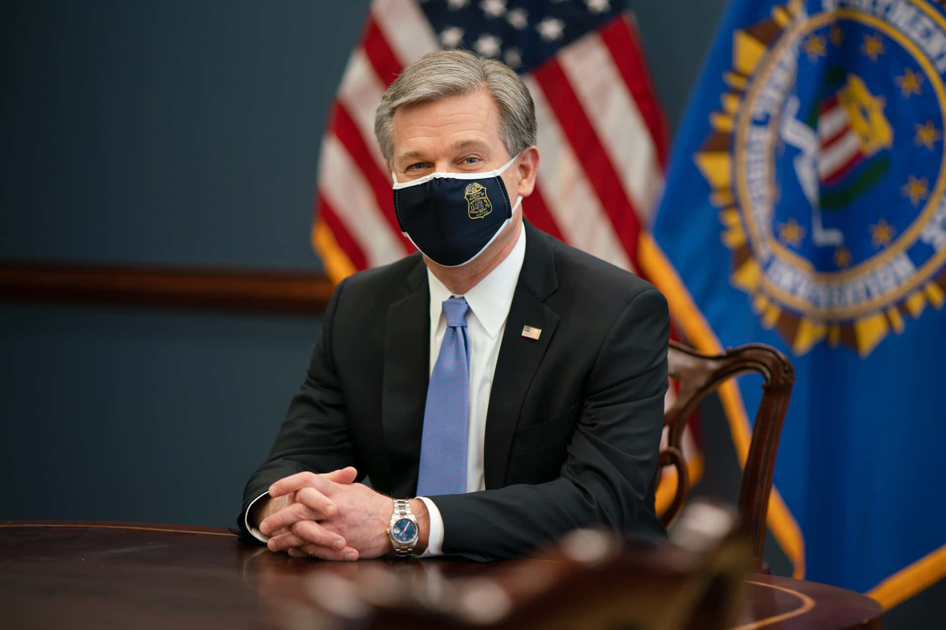 A Man In A Suit Sitting At A Desk Wearing A Face Mask Background