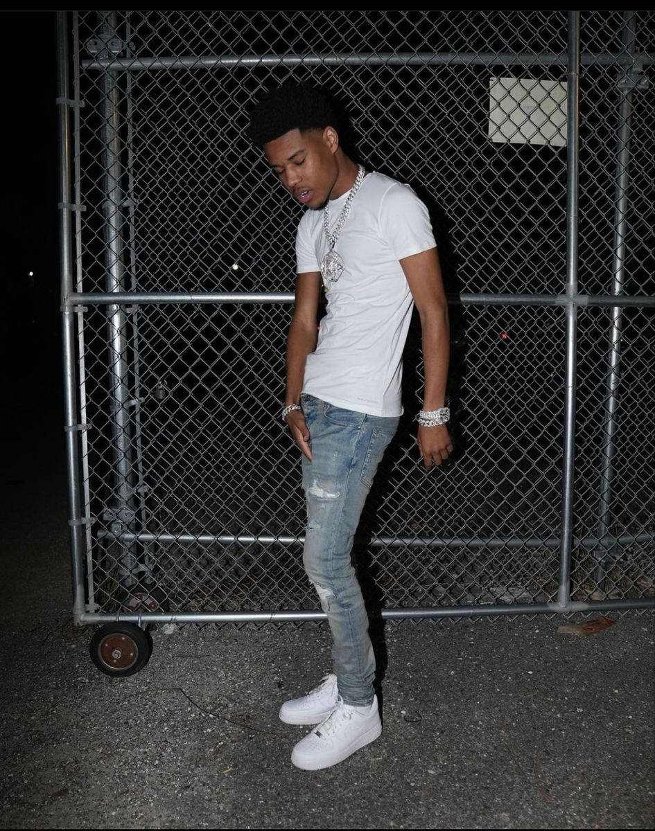 A Man In A White Shirt And Jeans Standing In Front Of A Fence Background