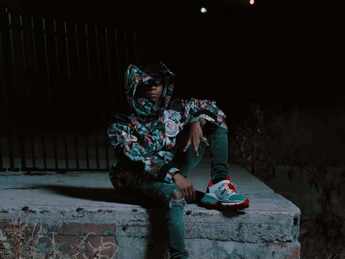 A Man Sitting On A Wall At Night Wearing A Hoodie And Sneakers Background