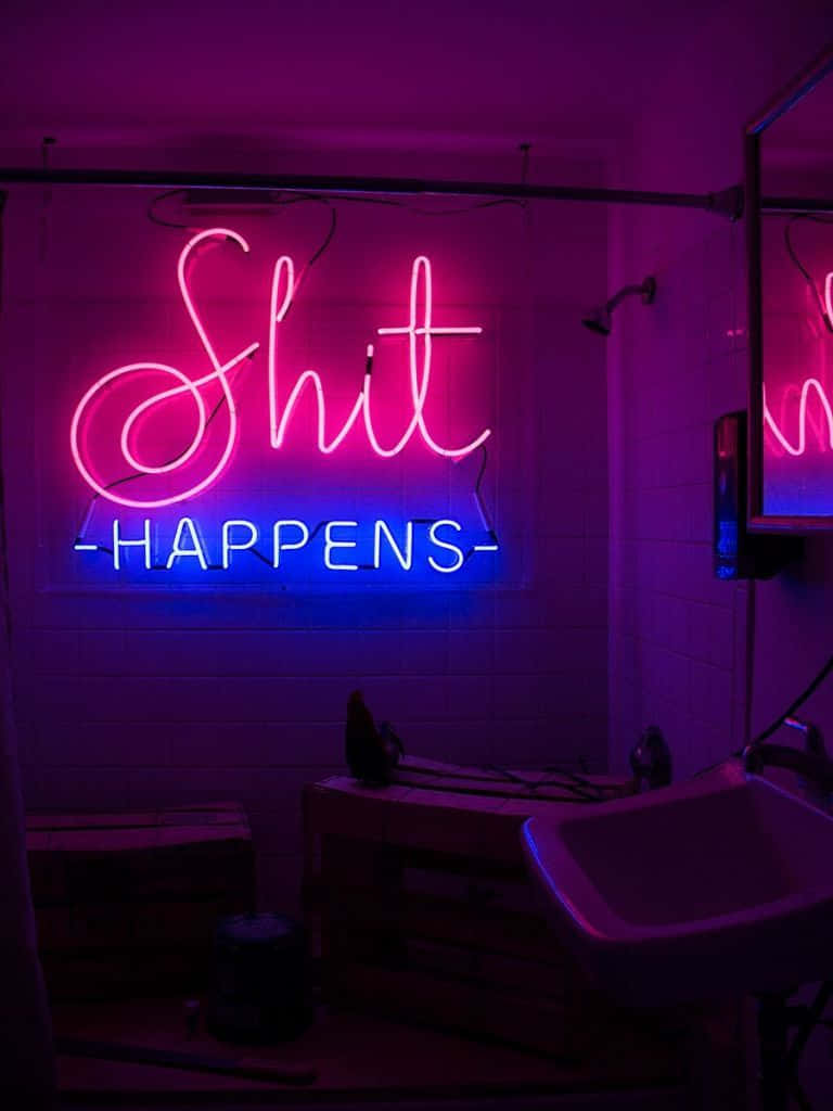 A Neon Sign That Says Shit Happens In A Bathroom Background