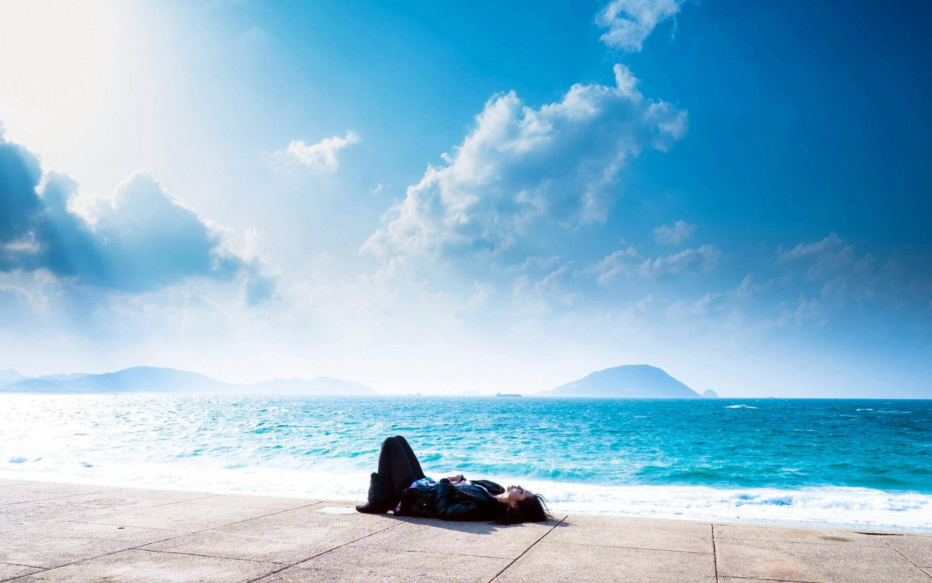 A Person Laying On A Bench Near The Ocean Background