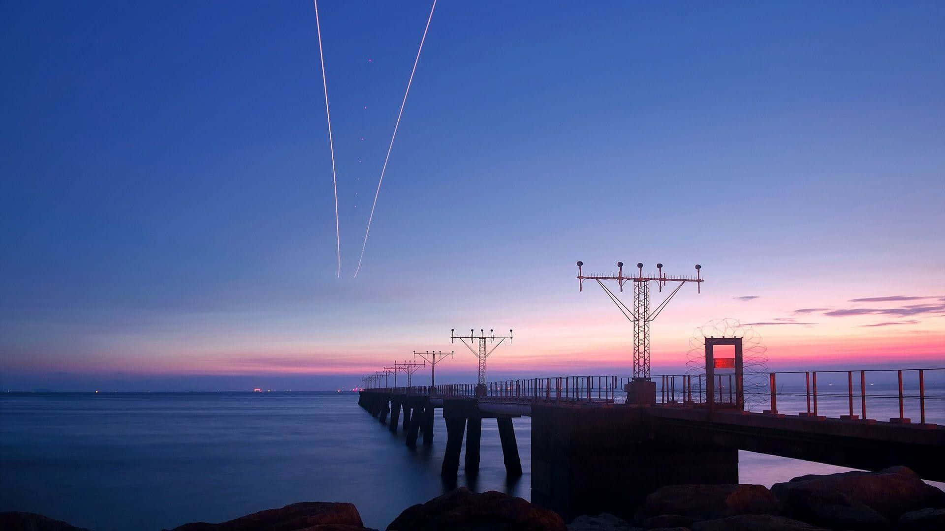 A Plane Flying Over A Pier At Sunset Background