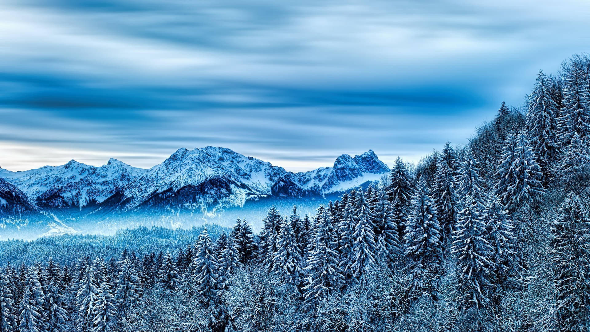 A Snowy Mountain Range With Trees And A Blue Sky Background