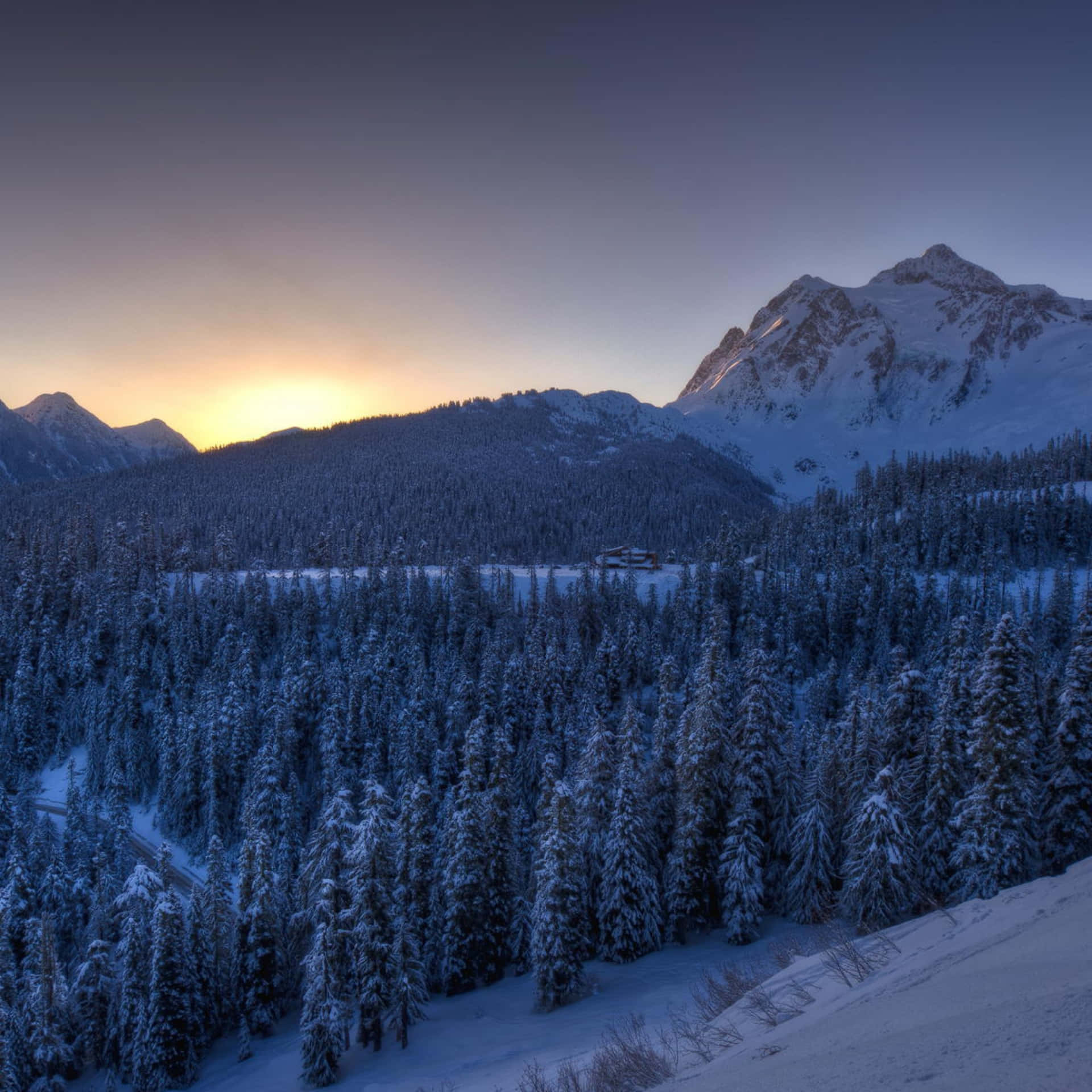 A Snowy Mountain Range With Trees And A Sun Setting Background