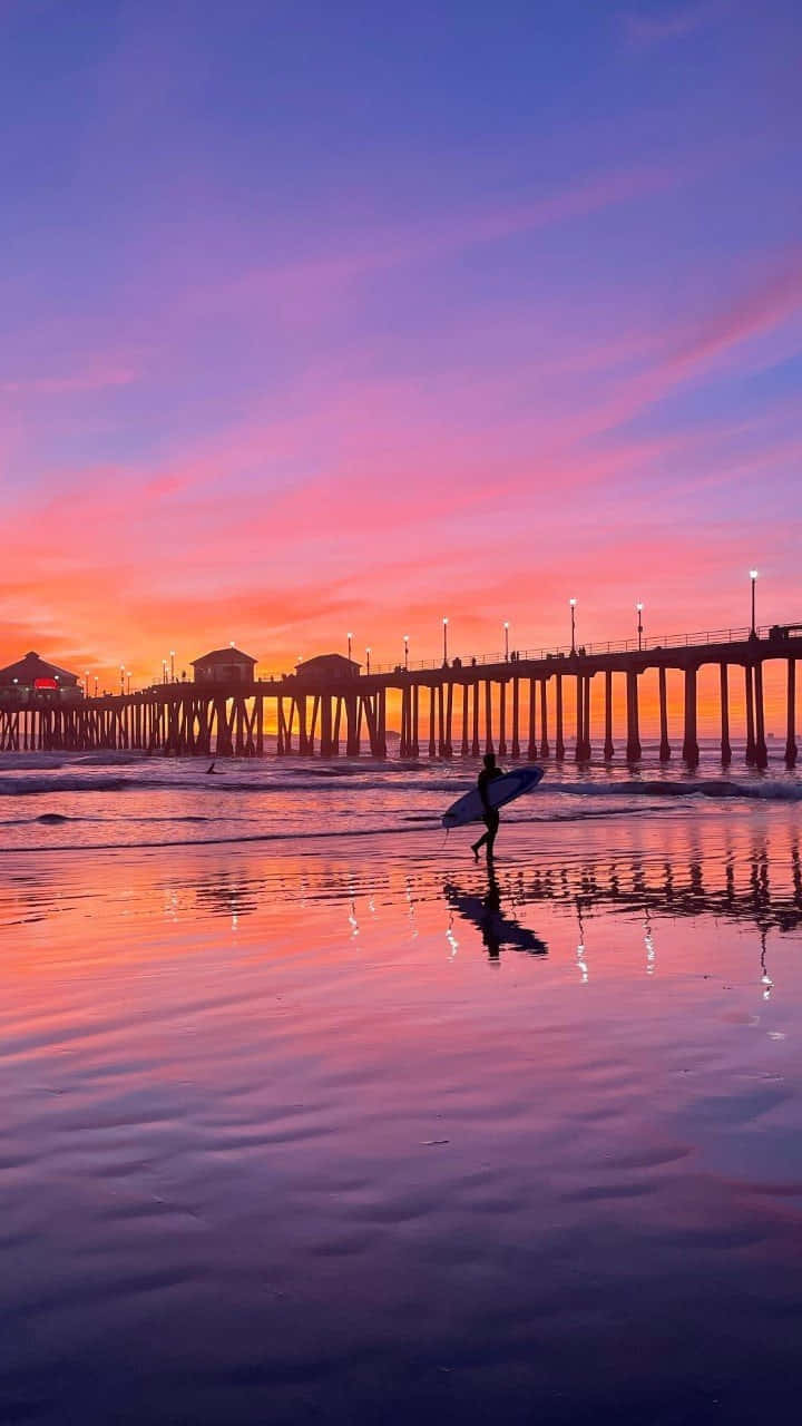 A Surfer Walks Along The Beach At Sunset Background