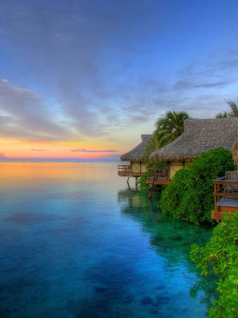 A Tropical Island With Thatched Huts And A Sunset Background