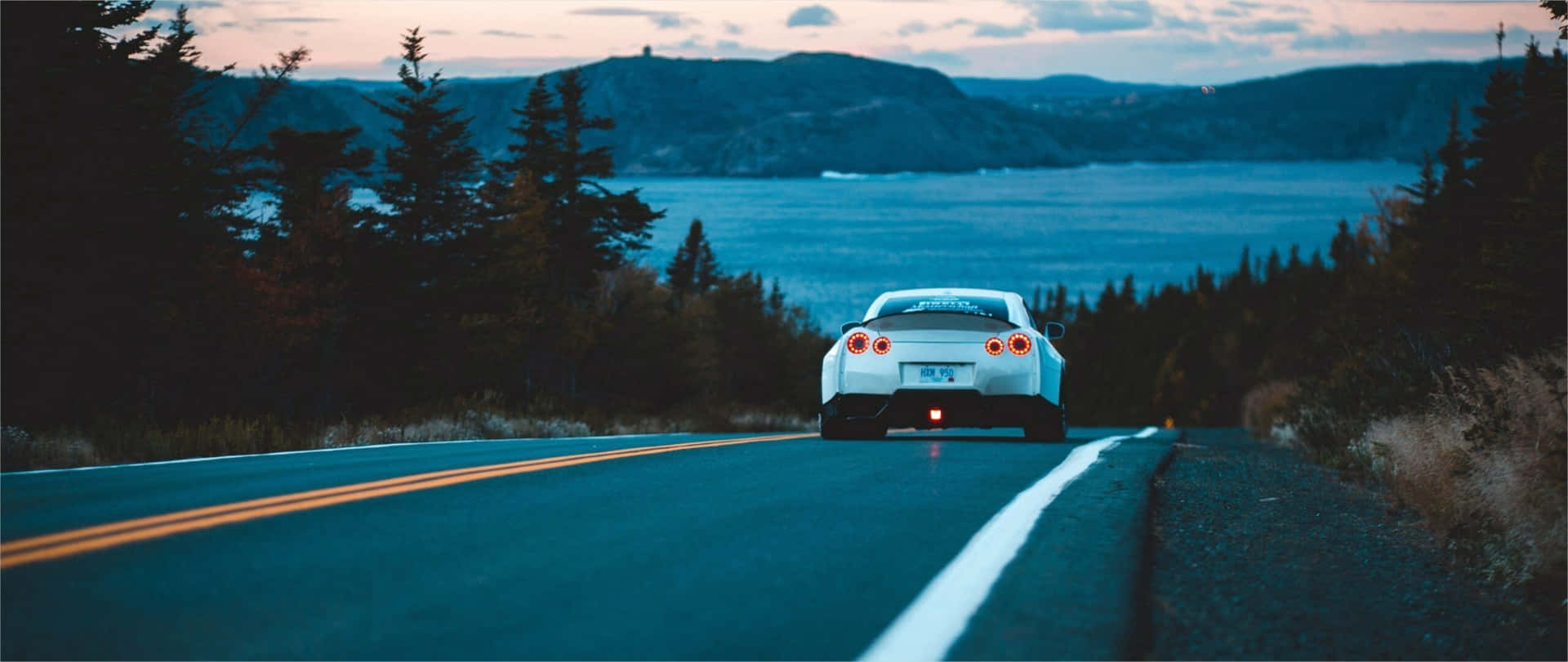 A White Car Driving Down A Road Near A Lake Background