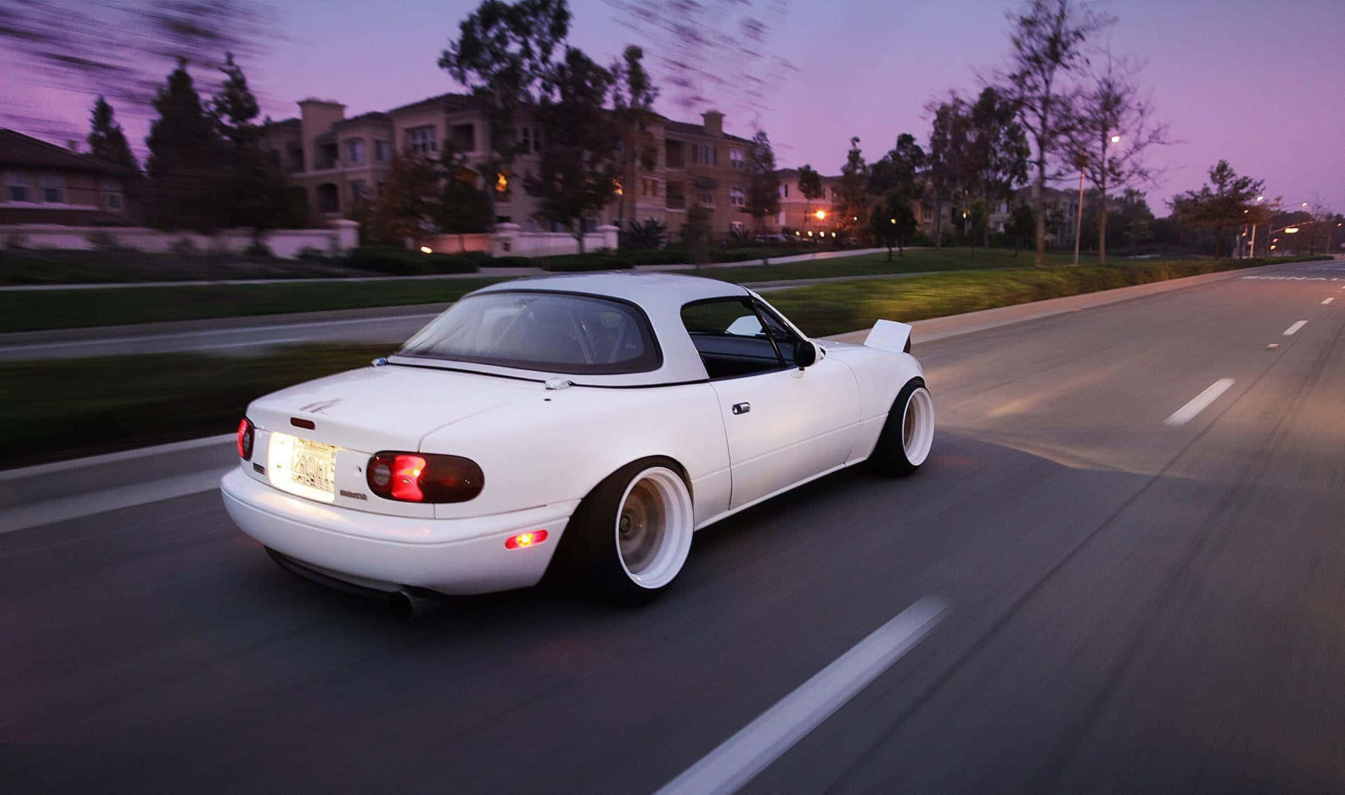 A White Sports Car Driving Down The Street At Dusk Background