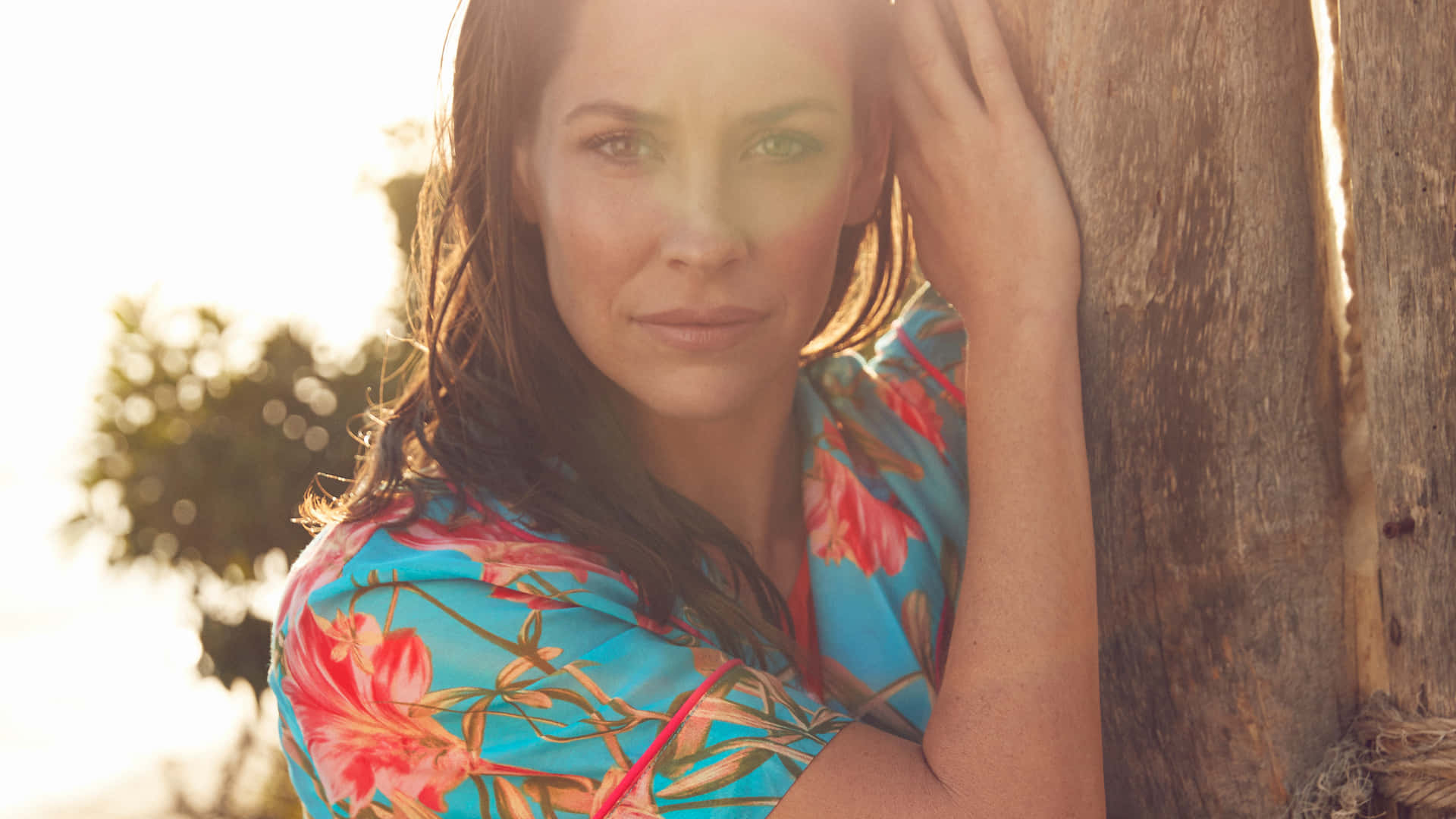 A Woman In A Floral Dress Leaning Against A Wooden Pole Background