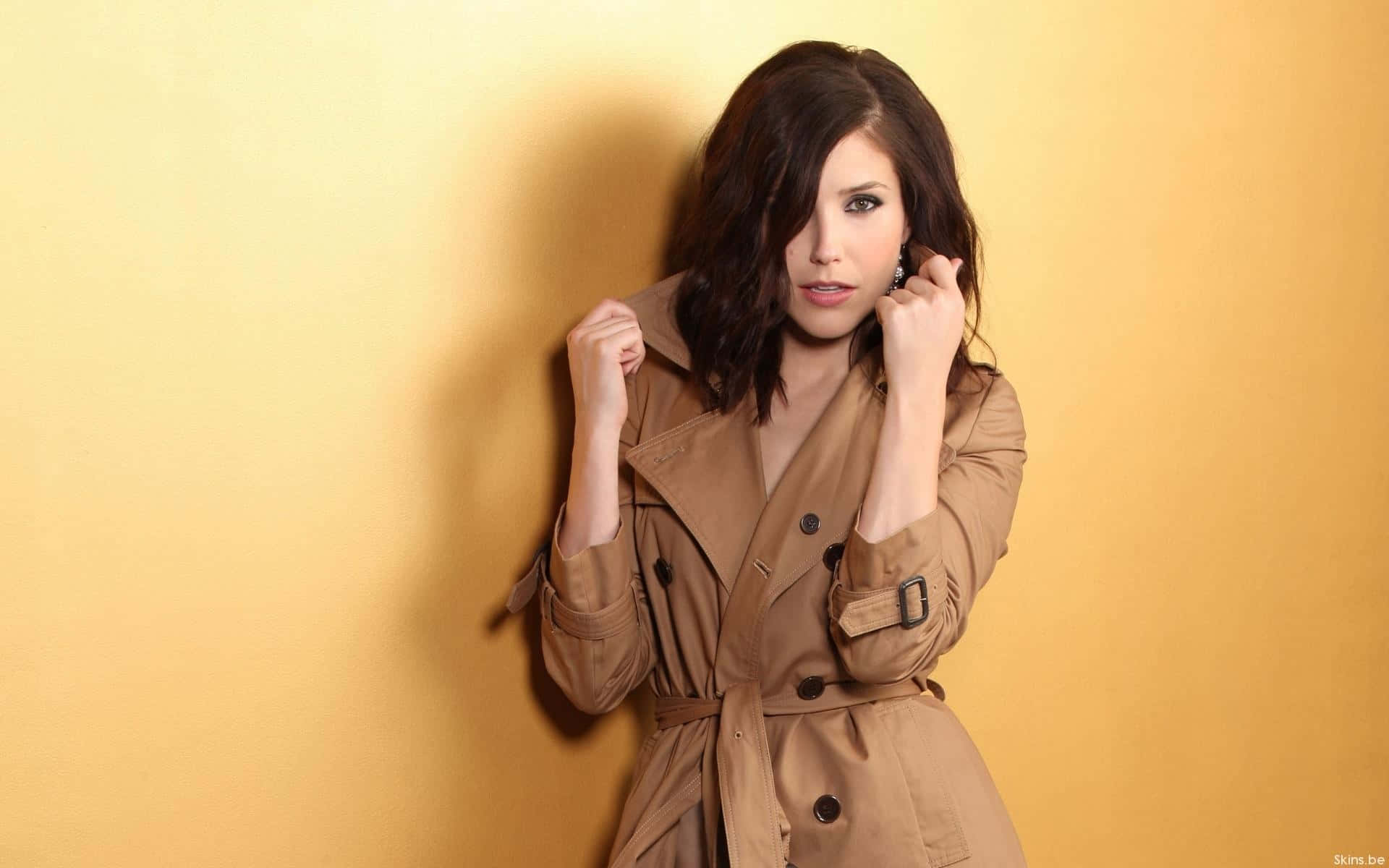 A Woman In A Trench Coat Leaning Against A Yellow Wall Background