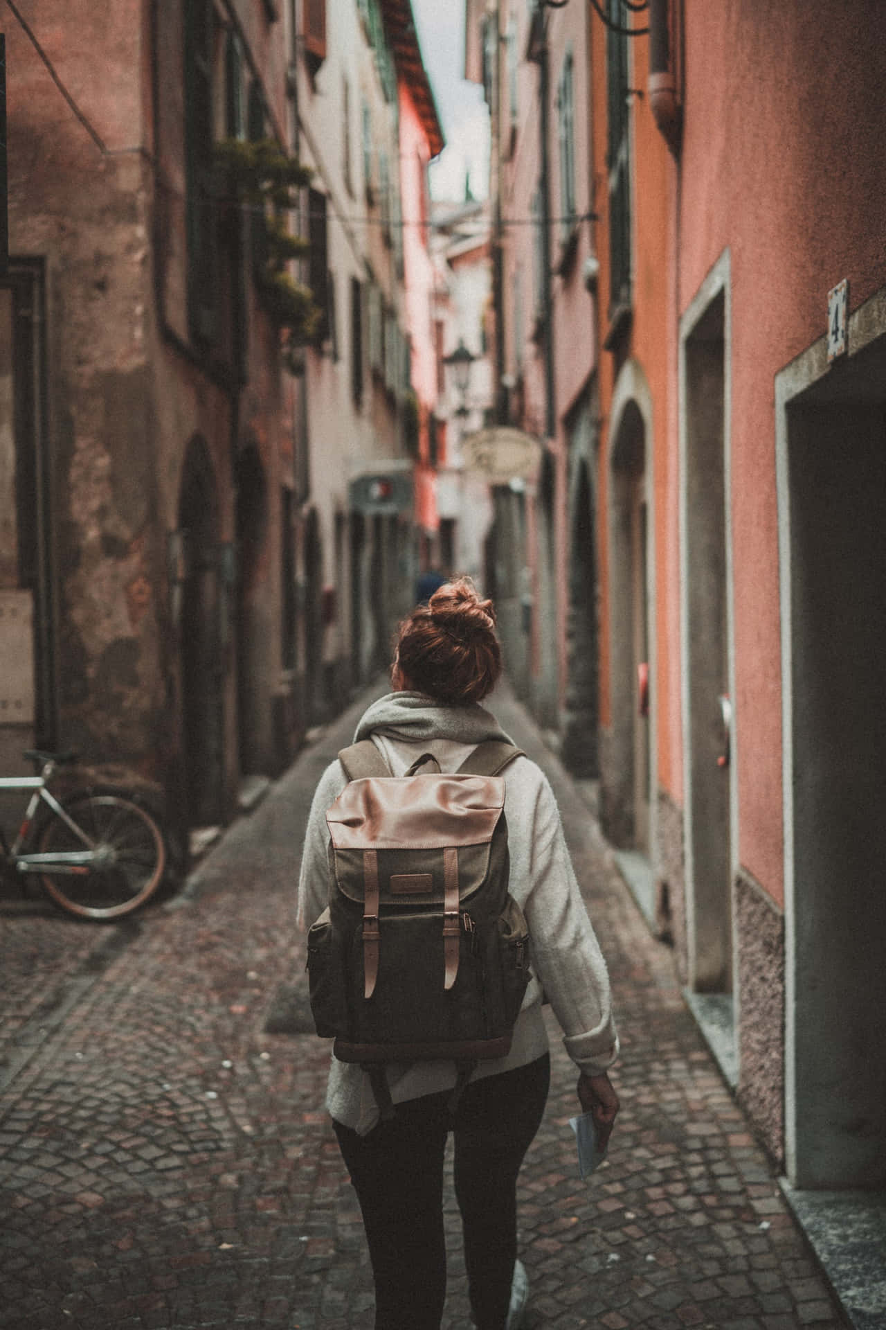 A Woman Walking Down A Narrow Street With A Backpack Background