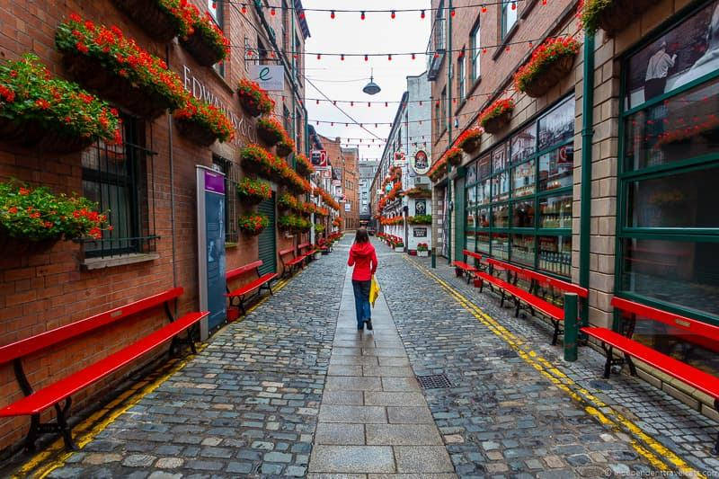 A Woman Walking Down A Narrow Street With Red Benches