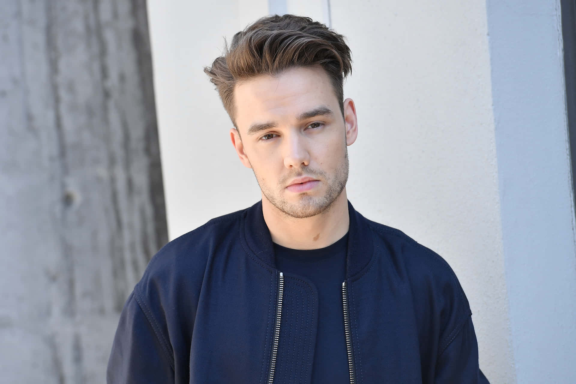 A Young Man In A Blue Jacket Leaning Against A Wall Background