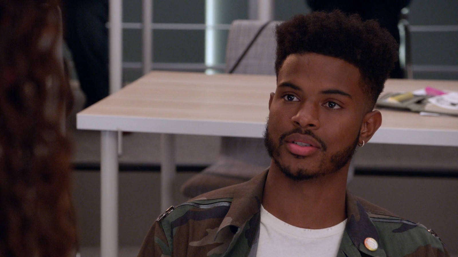 A Young Man In A Camouflage Jacket Is Sitting In An Office Background