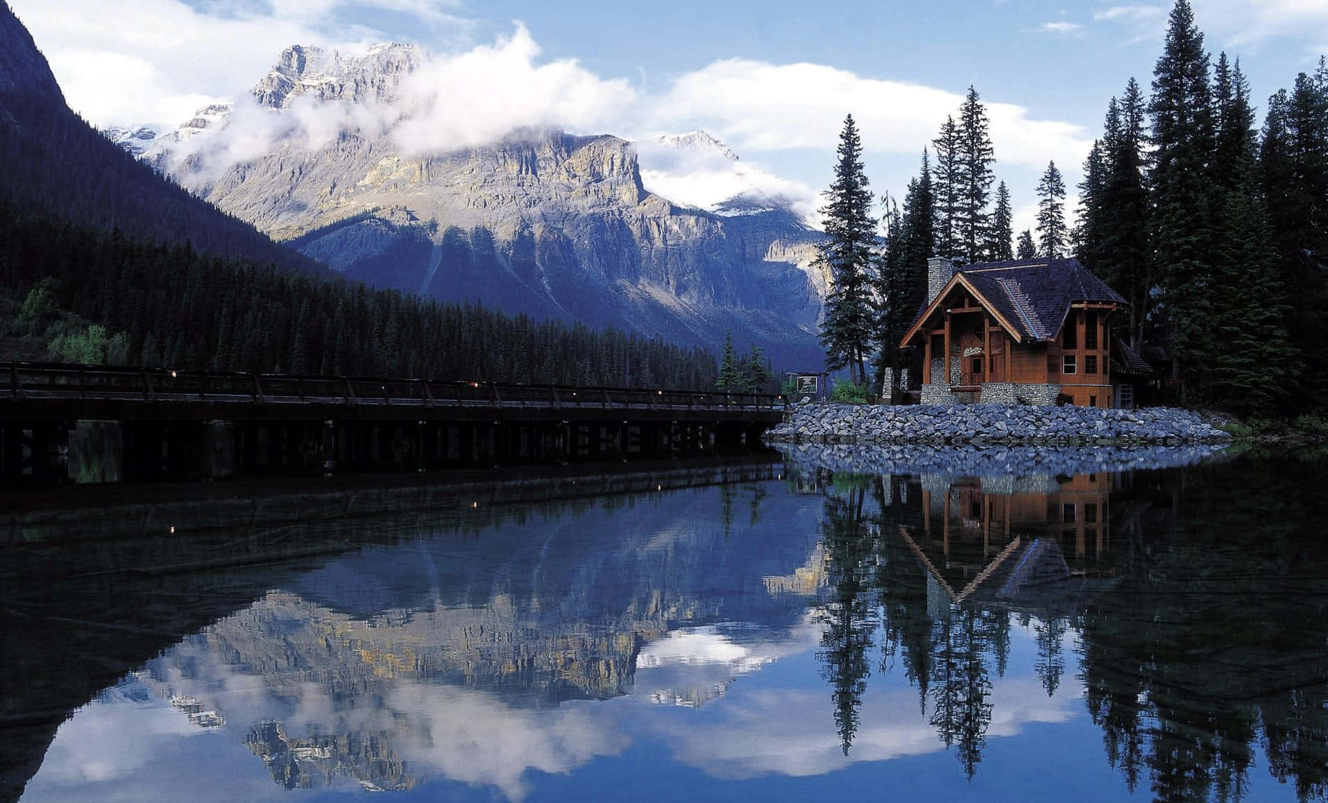 Cabin By A River Surrounded By Mountains And Nature Background