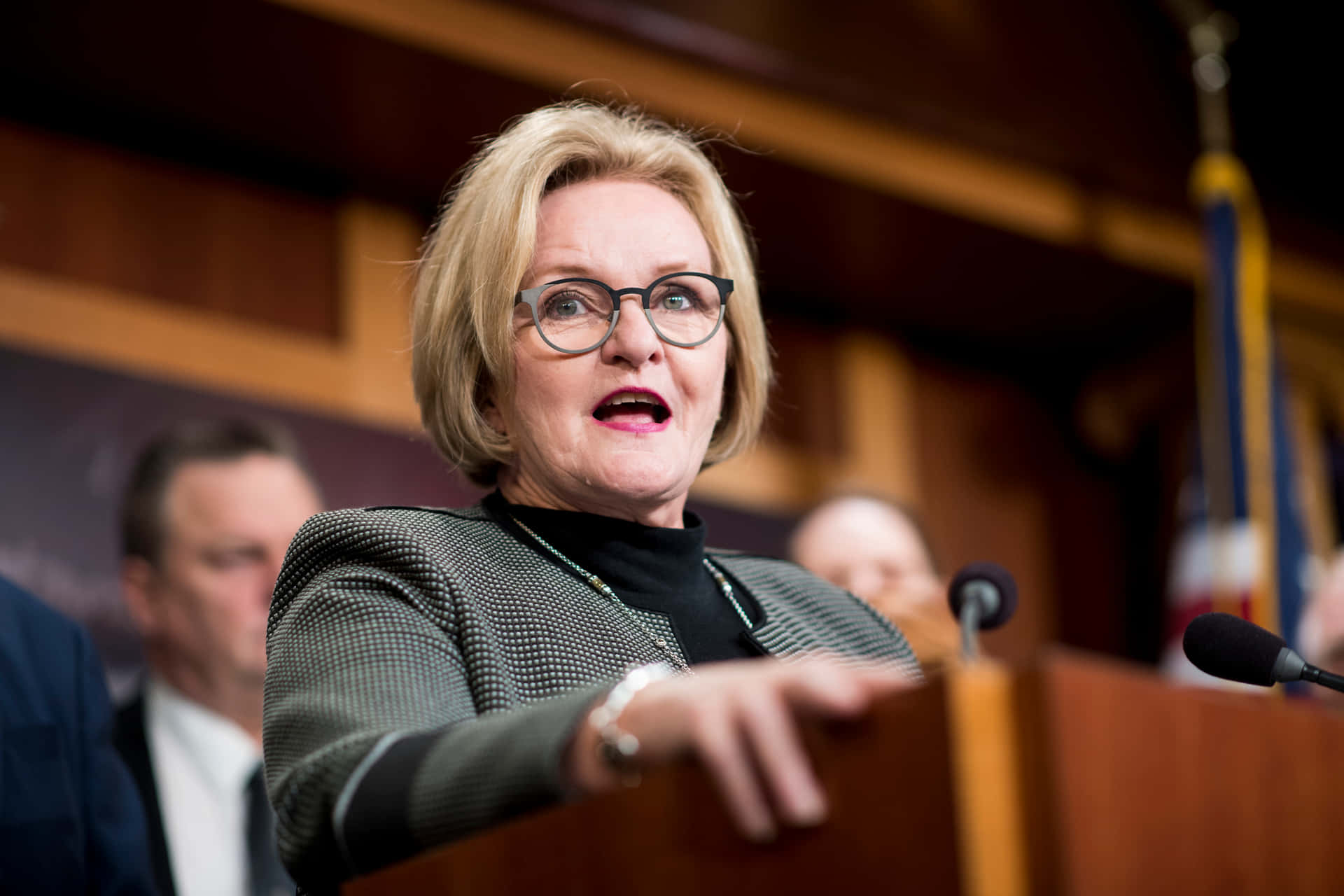 Claire Mccaskill Delivering A Speech On Stage With Fellow Congresspersons Background