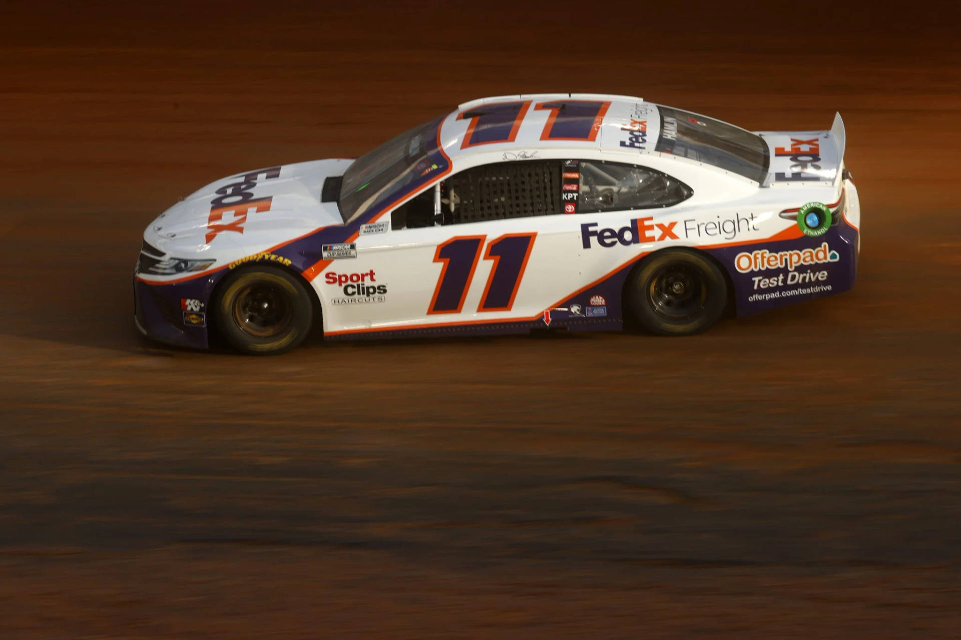 Denny Hamlin Skillfully Navigates Dirt Road In A High-speed Race Background