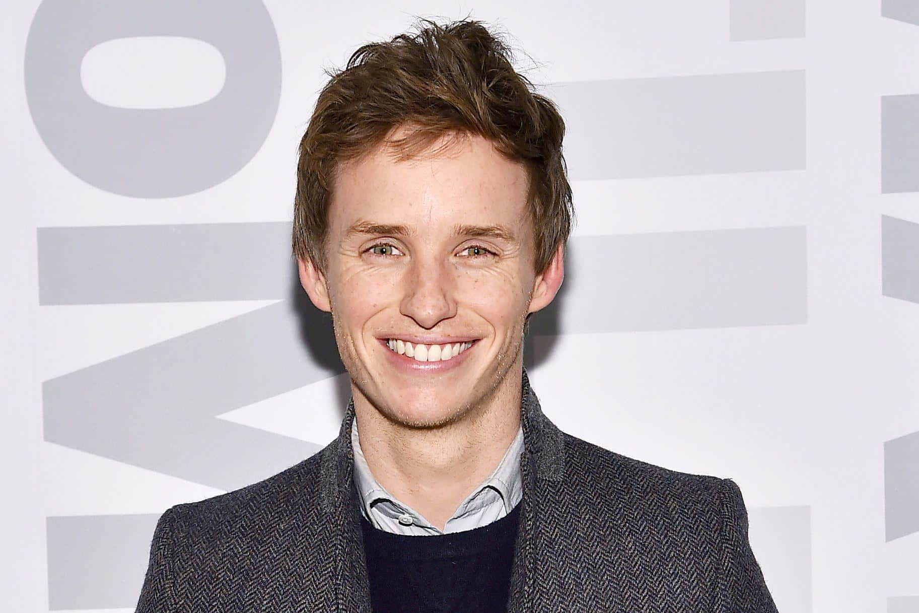 Eddie Redmayne Smiling In A Black Suit Against A Maroon Background Background