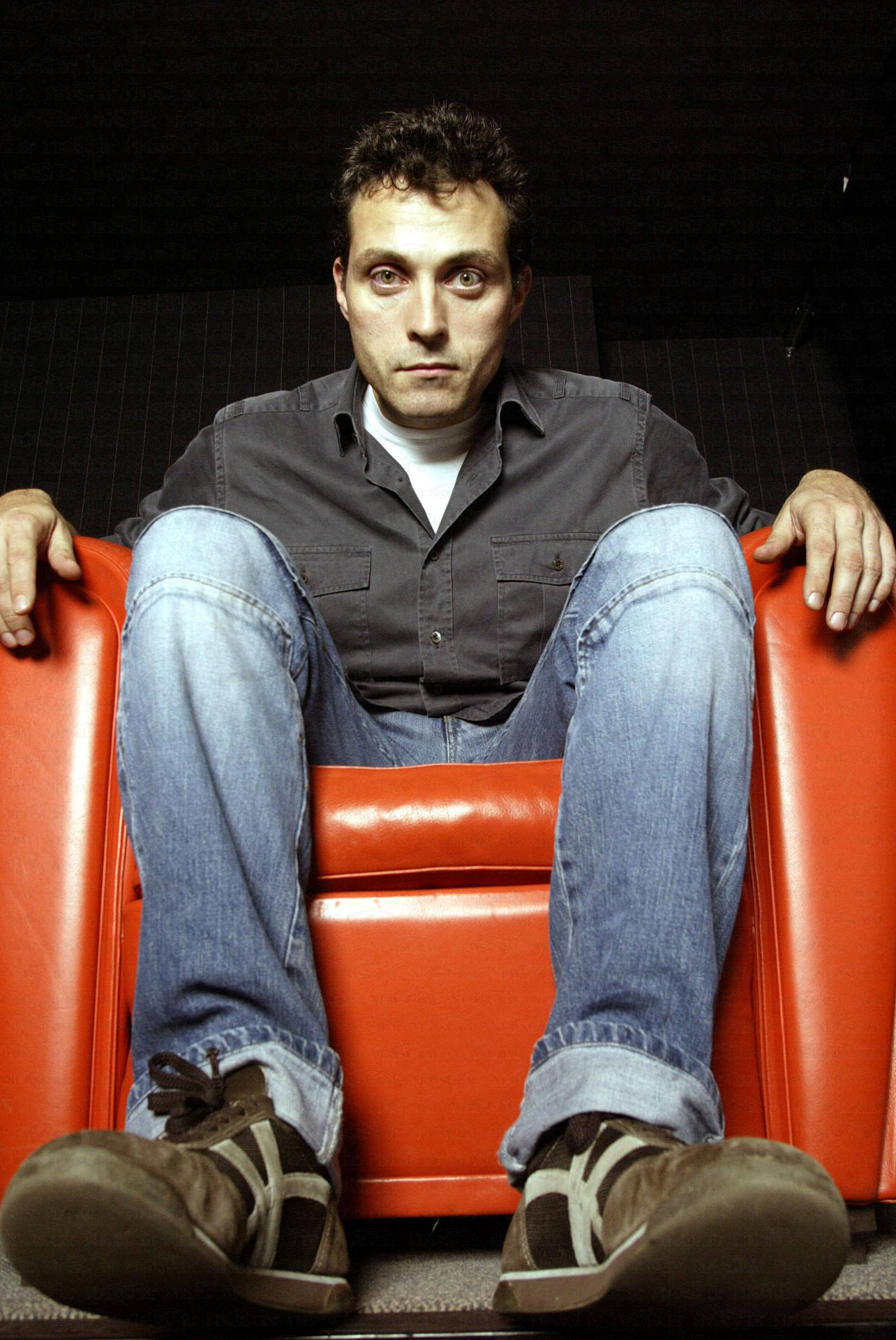 Eerie Rufus Sewell Sitting On An Orange Recliner Background