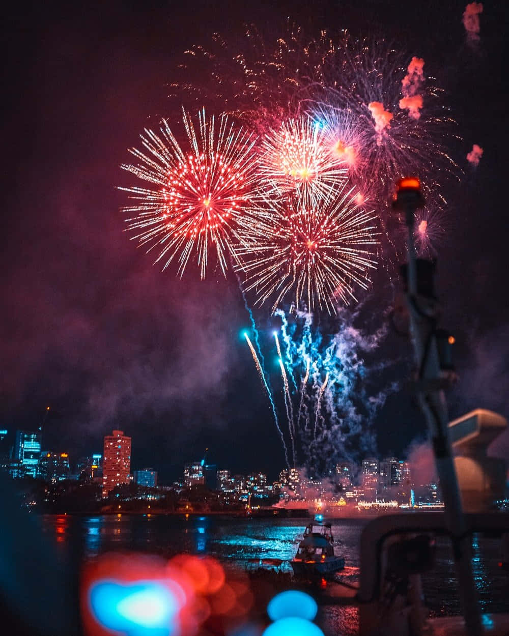 Fireworks Over A City With A Boat In The Background Background
