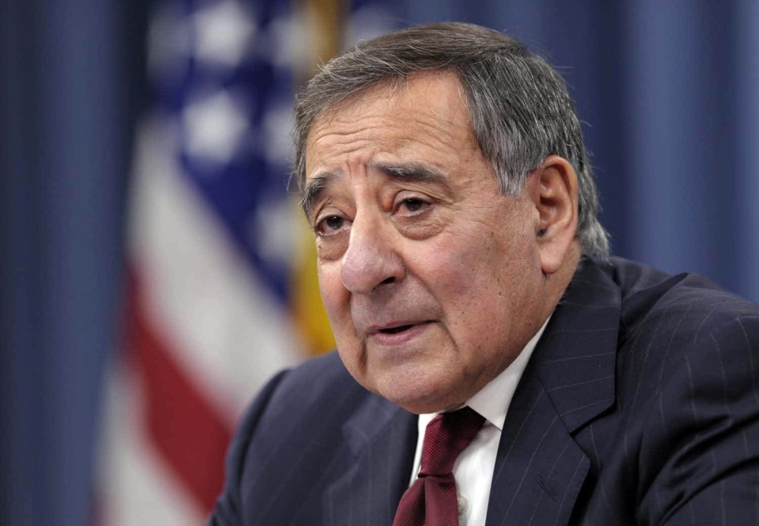 Former Secretary Of Defense Leon Panetta Stands In Front Of A Blurred American Flag. Background