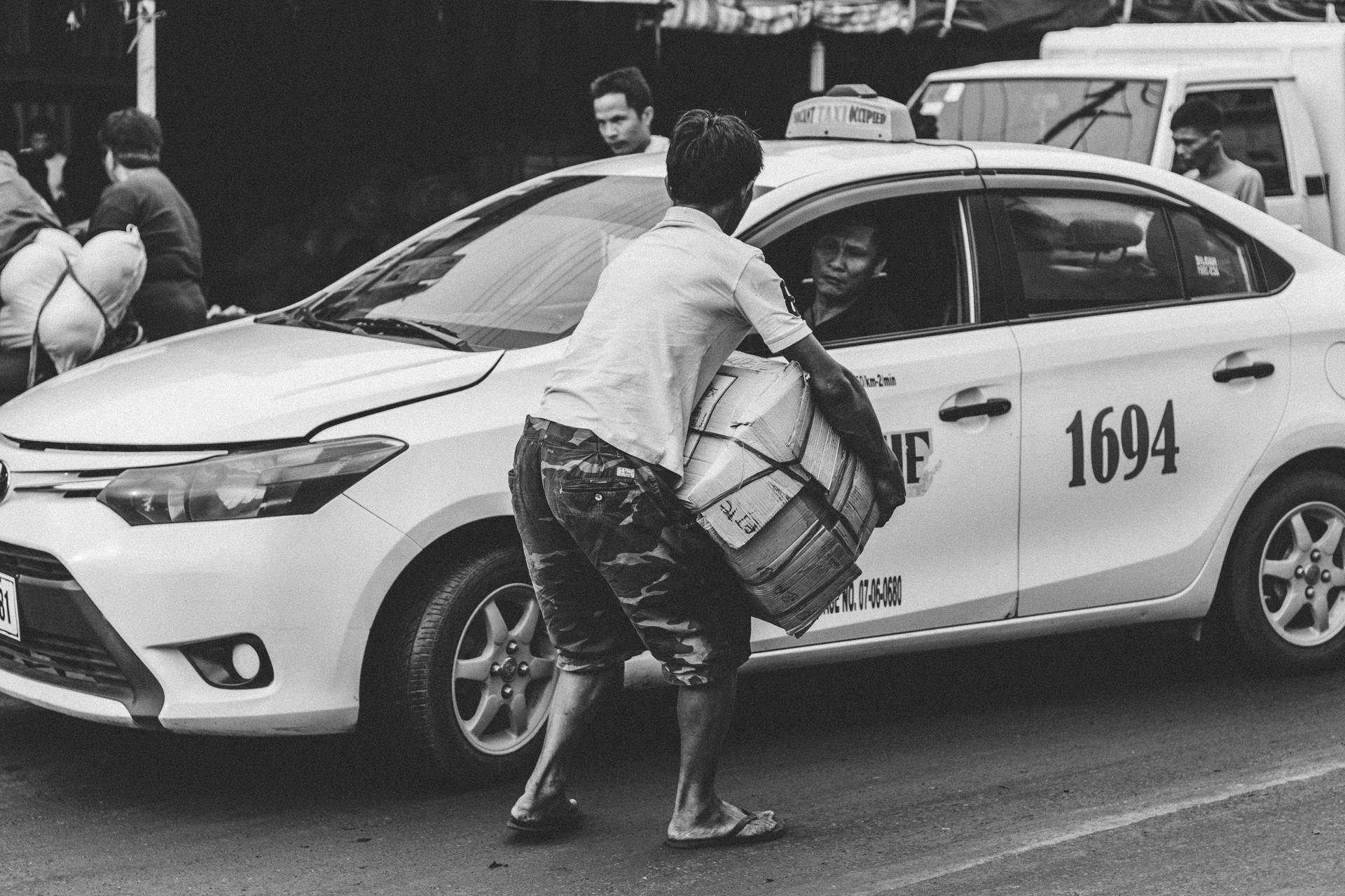 Greyscale Photography Of Man Carrying Cardboard Box Beside Taxi Background
