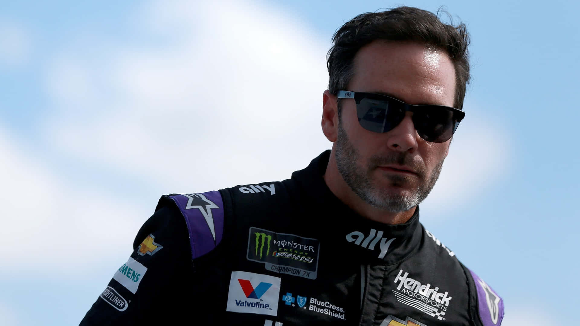 Jimmie Johnson In His Racing Gear With His Car On The Track Background