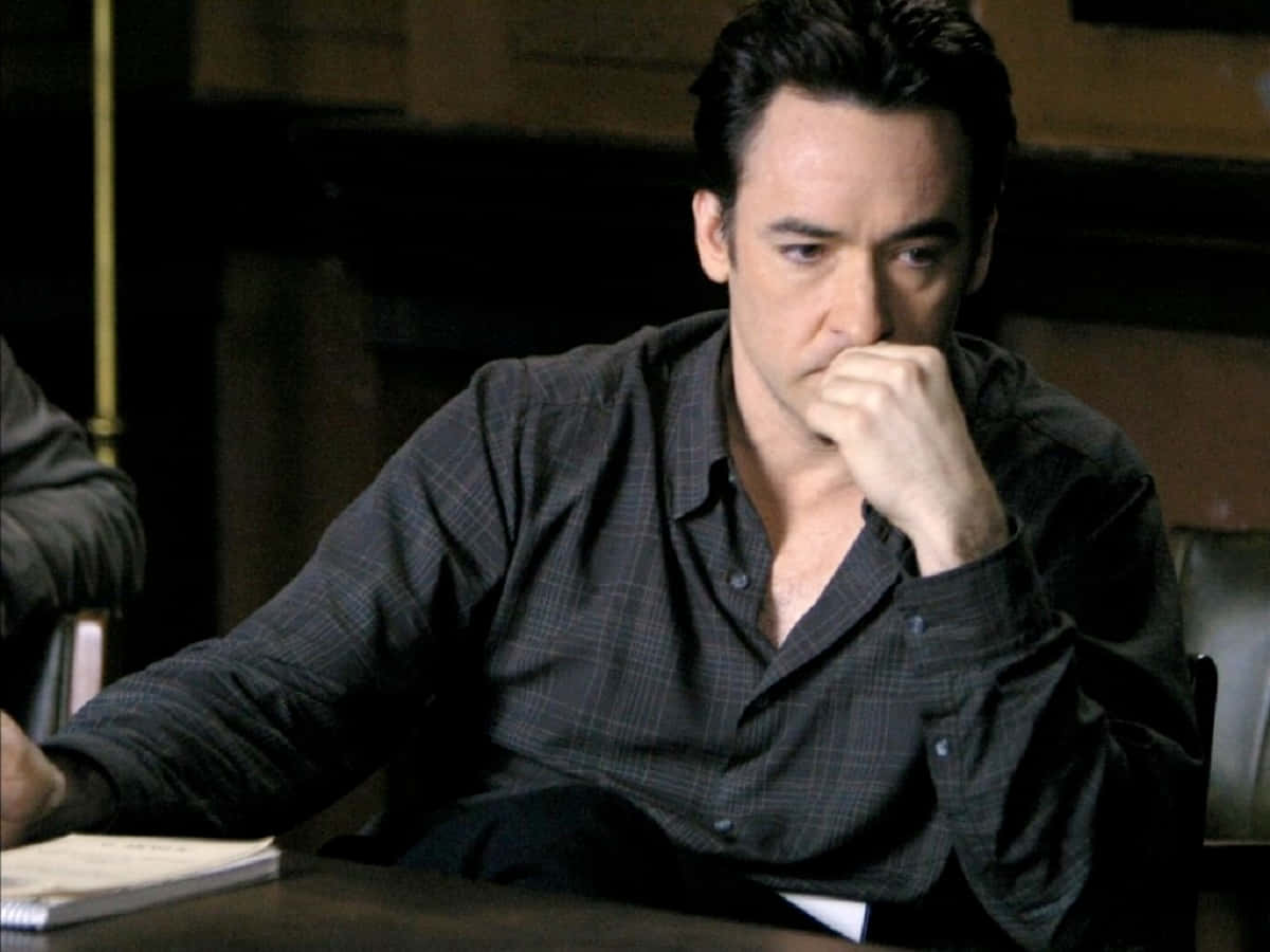 John Cusack Strikes A Casual Pose In Black Attire Against A Blurred Cityscape Background