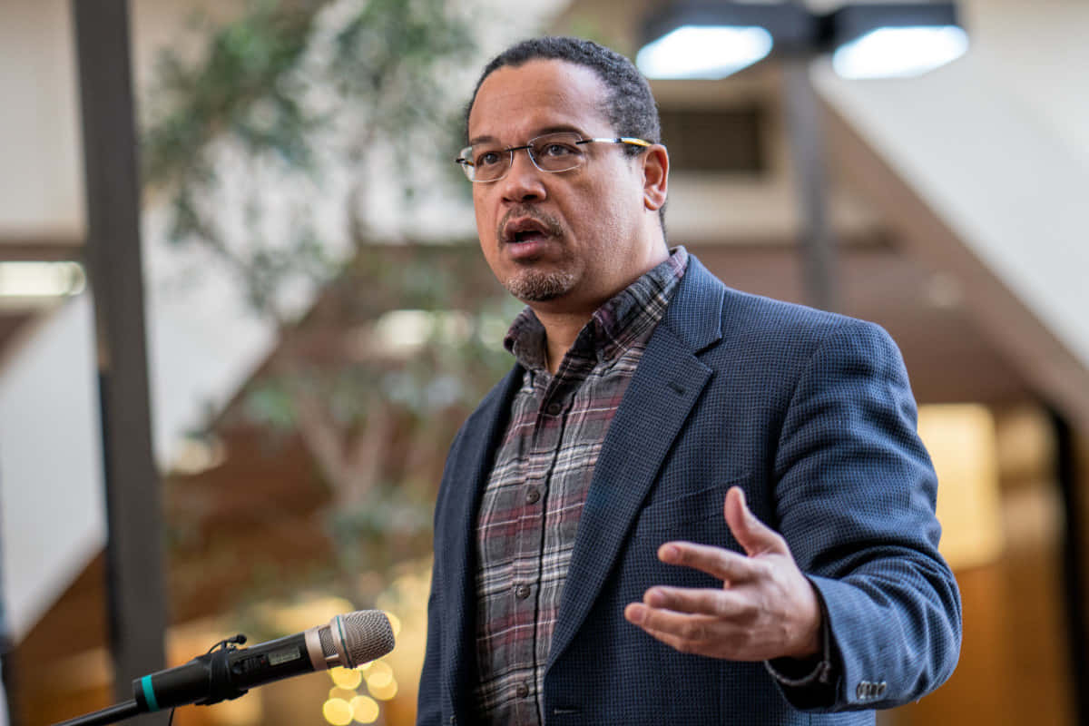 Keith Ellison Looking Professional In Plaid Shirt And Blazer Background