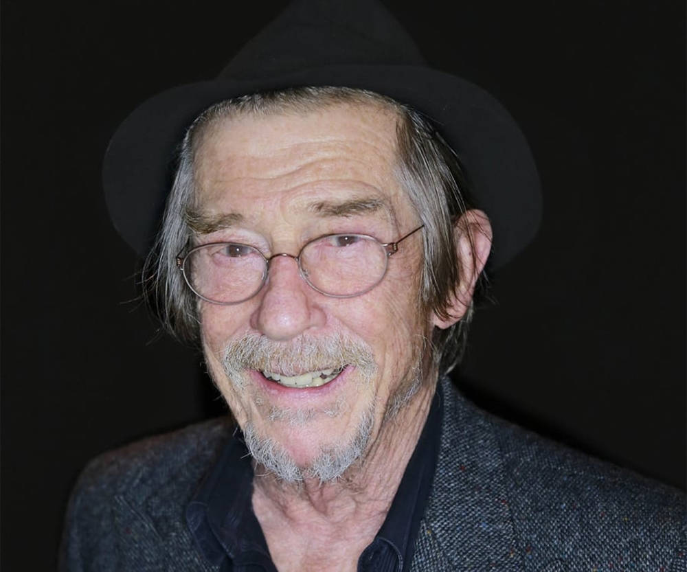 Legendary Actor John Hurt In Hat And Glasses Against A Black Background