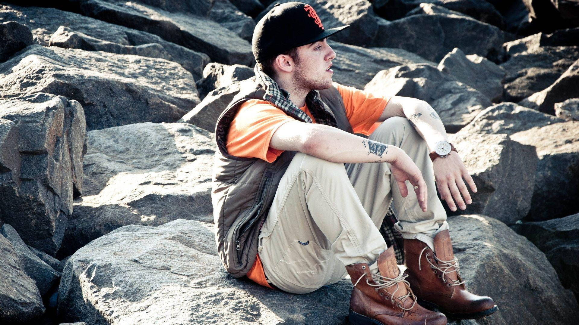 Mac Miller Sitting On Rocks Background