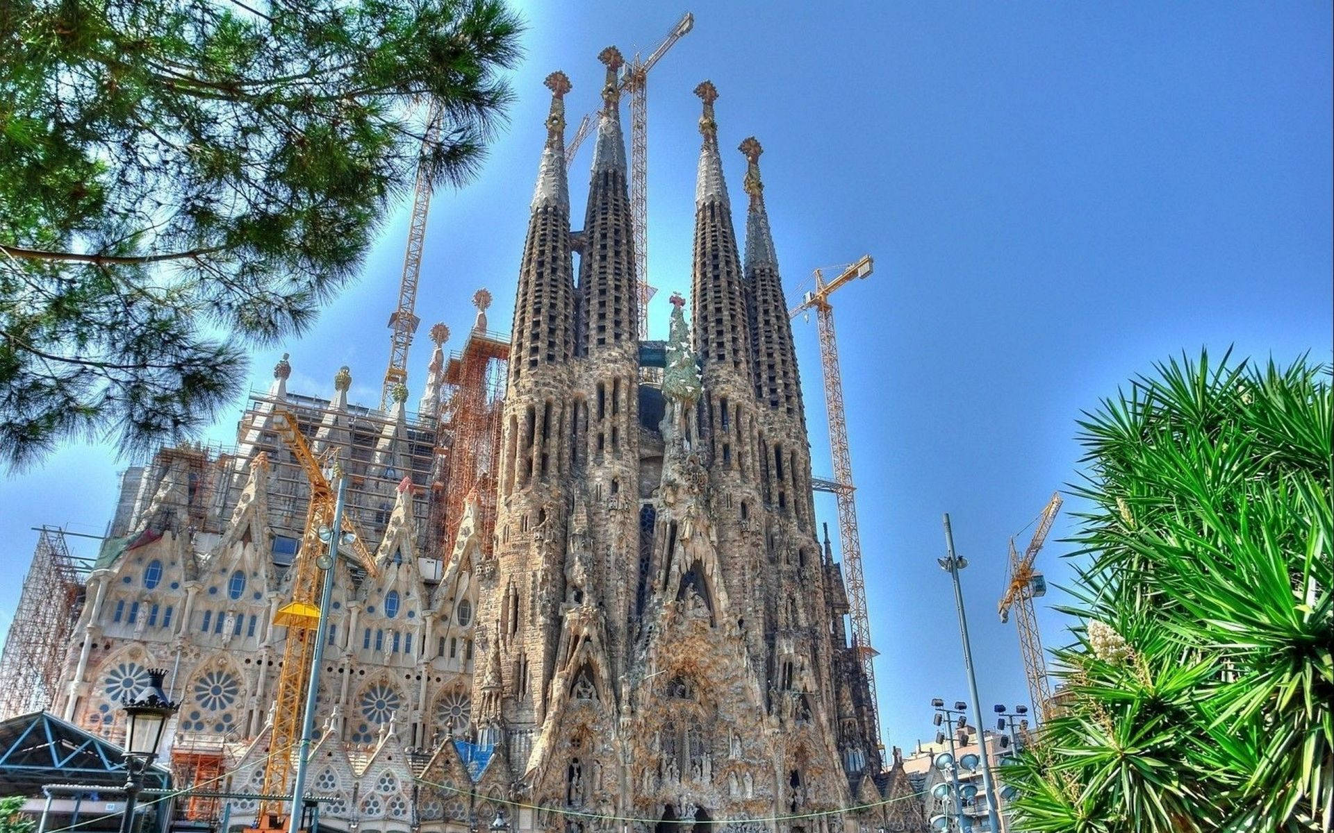 Majestic Sagrada Familia Surrounded By Lush Trees And Industrial Cranes Background