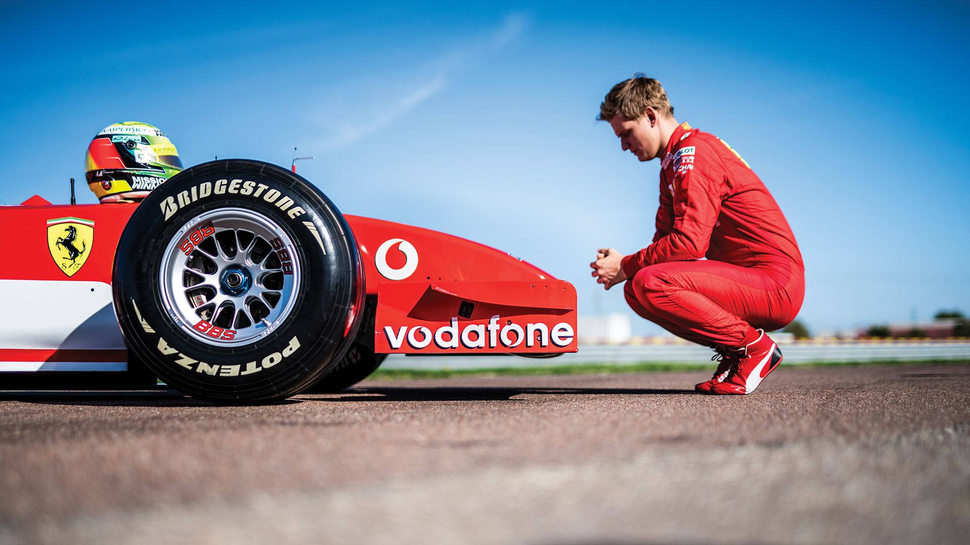 Mick Schumacher Crouching Beside A Car Background